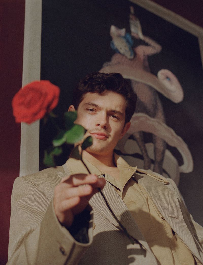 Young man in a beige suit holds out a red rose, portrait indoors with a painting background, medium shot, soft lighting, direct gaze.