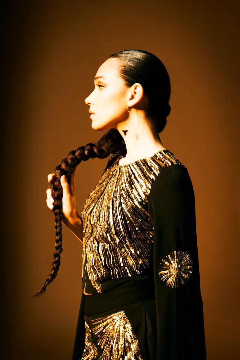 Woman in profile with long braid and dark dress with gold embellishments, studio lighting against a brown backdrop. Fashion and beauty shot.