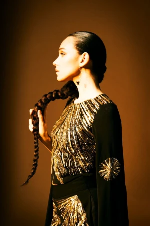 Woman in profile with long braid and dark dress with gold embellishments, studio lighting against a brown backdrop. Fashion and beauty shot.
