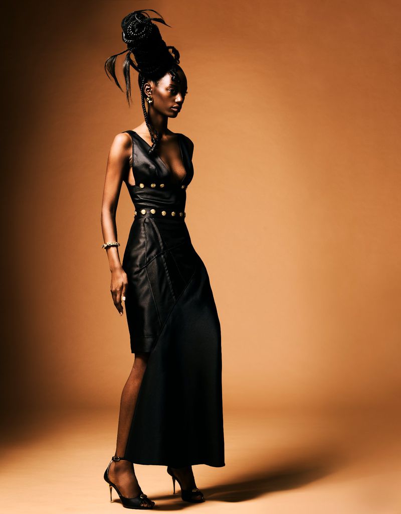 Portrait of a Black female model in a stylish black dress with gold accents posing in a studio with a tan background and dramatic lighting.