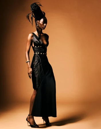 Portrait of a Black female model in a stylish black dress with gold accents posing in a studio with a tan background and dramatic lighting.