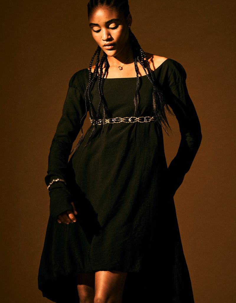 Portrait of a Black woman in a black dress with long sleeves, styled with braids and a silver chain belt against a brown backdrop.