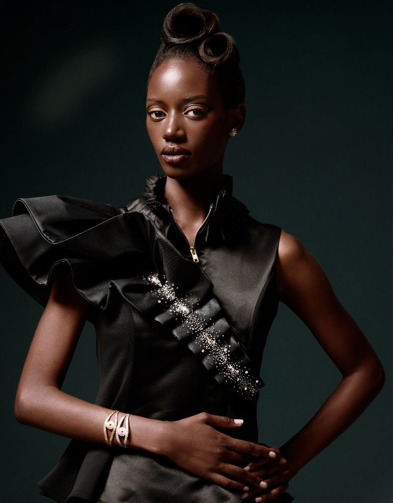 Close-up fashion portrait of a Black woman with elaborate hair, jewelry, and a ruffled black dress against a dark teal background.