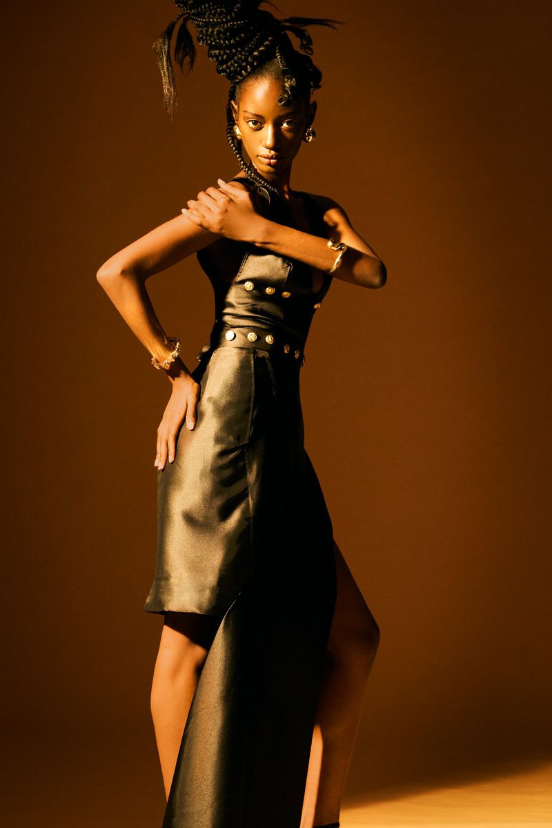 Fashionable black female model in a dark dress with buttons, bracelets and braided hair, posing in a warm, studio environment.