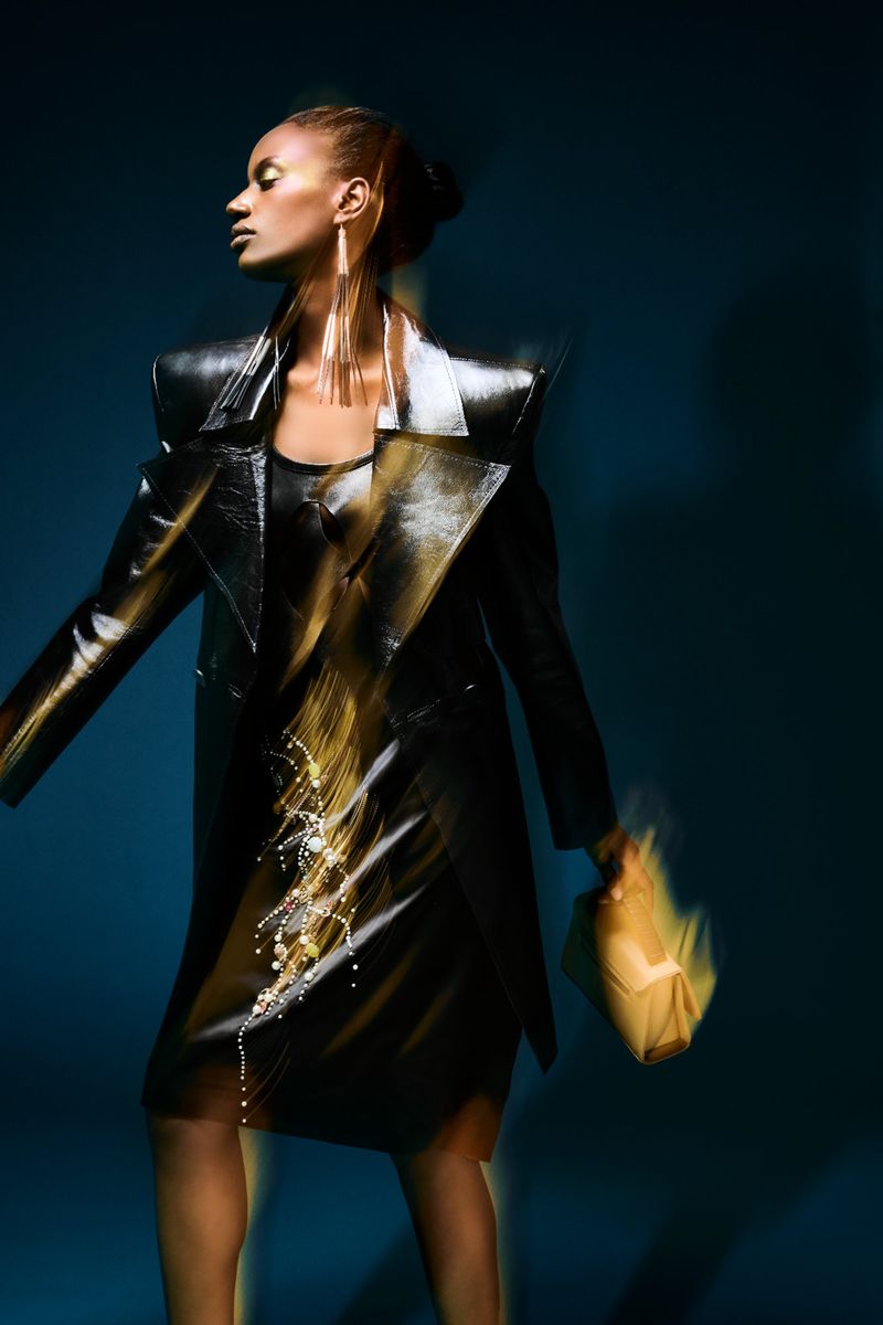 A woman in a dark coat and dress, wearing long earrings, poses against a blue background. She is holding a bag with blurred motion.