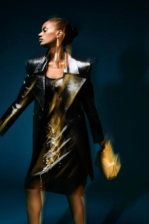 A woman in a dark coat and dress, wearing long earrings, poses against a blue background. She is holding a bag with blurred motion.
