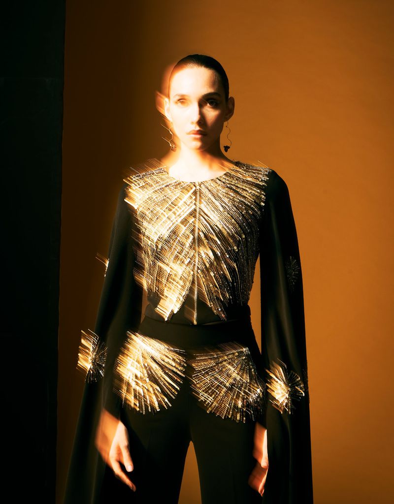 A fashionable adult female model poses in a black top and pants outfit with gold embellishments against a yellow-gold gradient background.