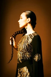 Side profile of a woman holding a long braided ponytail, wearing a black top with gold sequin embellishments against a warm background.