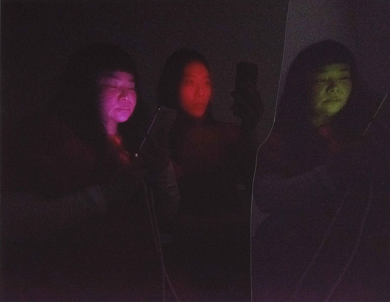 Three mirrored reflections of an Asian woman in dark surroundings illuminated by coloured phone screen light.