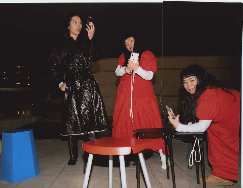 Three Asian women in stylish outfits are posing on a rooftop at night, each holding a smartphone, with modern stools nearby.