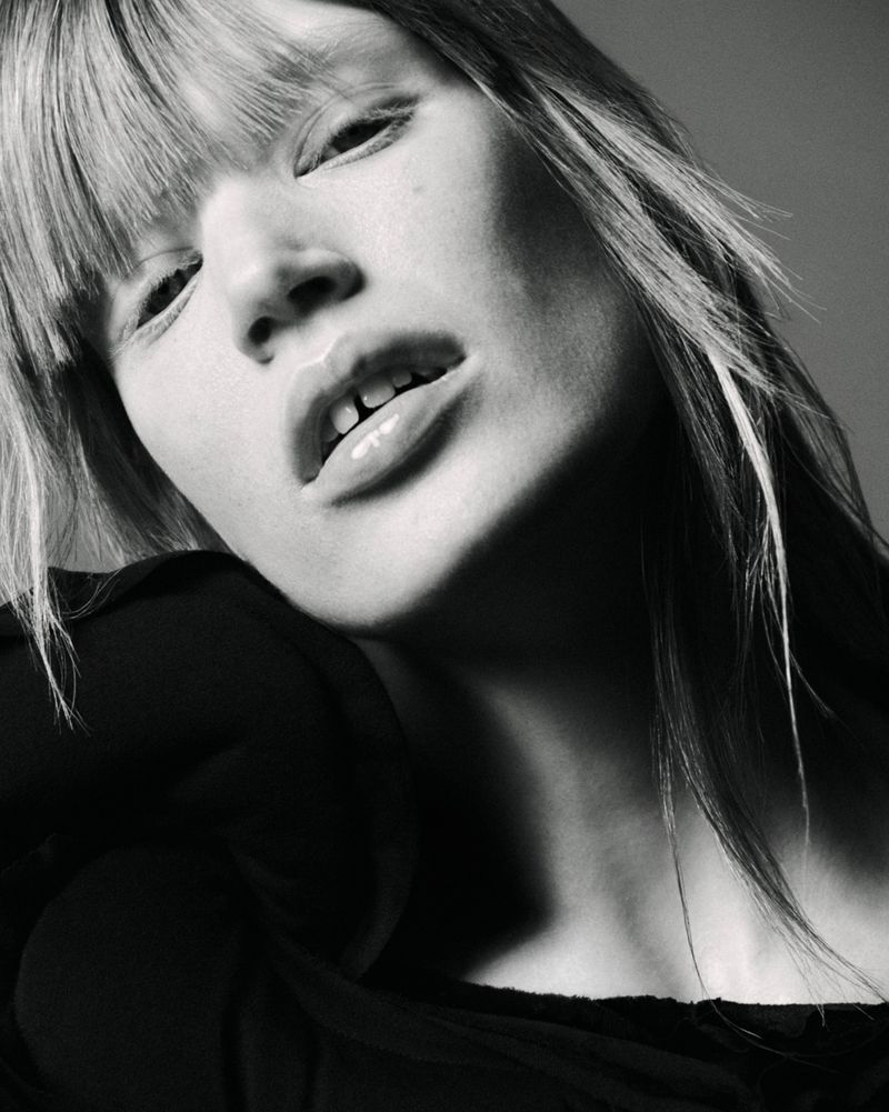 Close-up black and white portrait of a model with bangs wearing a dark top against a neutral studio background.