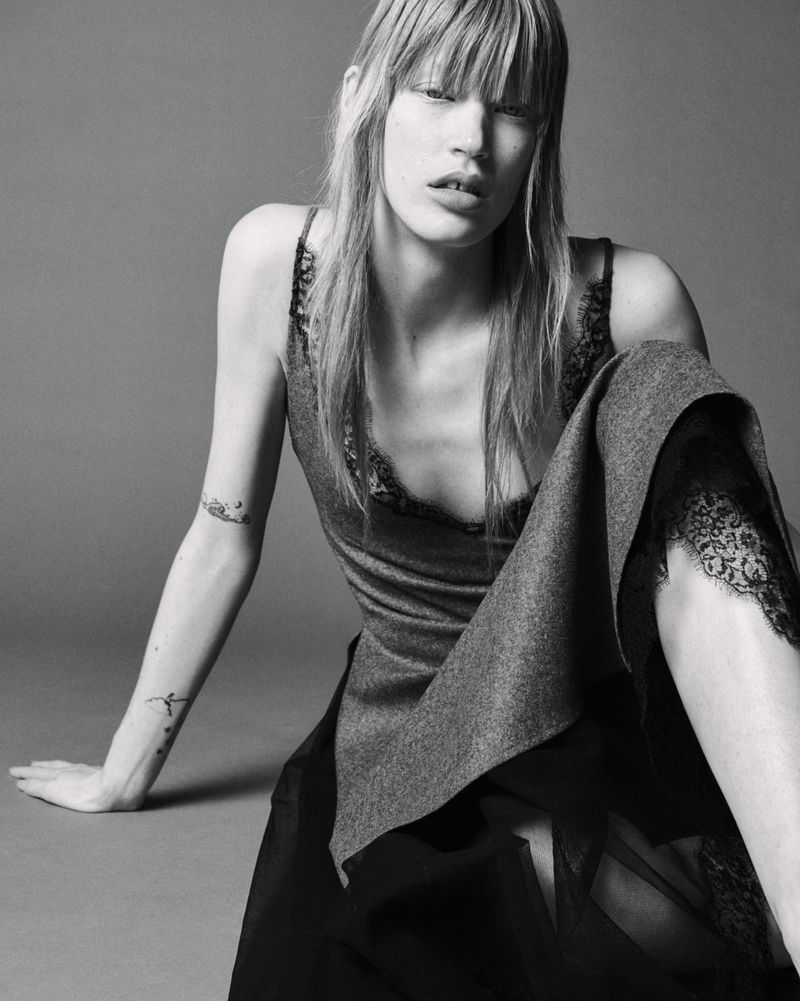 Model with long blonde hair and bangs wearing a lace-trimmed slip top and skirt, seated against a plain grey studio backdrop.