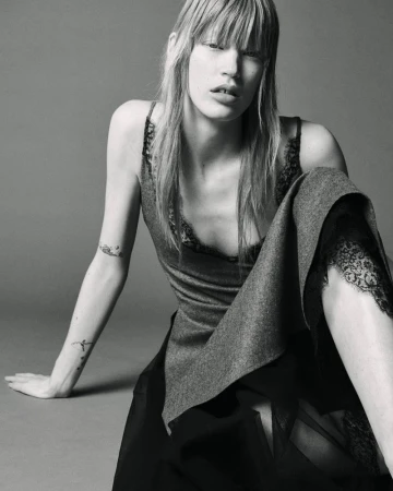 Model with long blonde hair and bangs wearing a lace-trimmed slip top and skirt, seated against a plain grey studio backdrop.