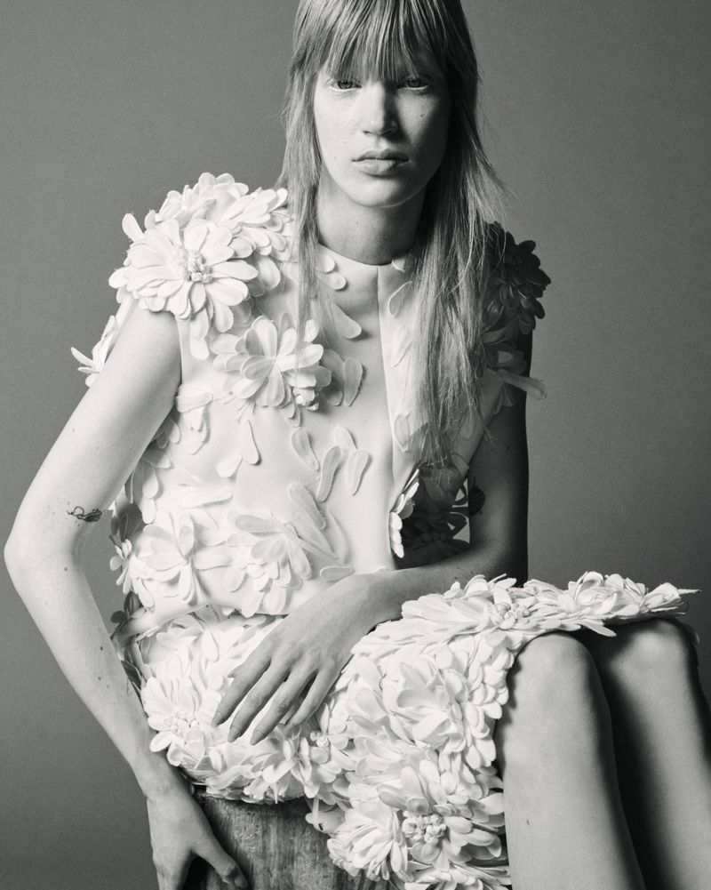 Model Jacqui Hooper in a white textured floral appliqué dress seated against a grey studio backdrop.