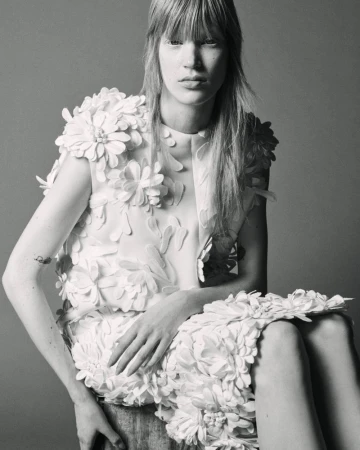 Model Jacqui Hooper in a white textured floral appliqué dress seated against a grey studio backdrop.