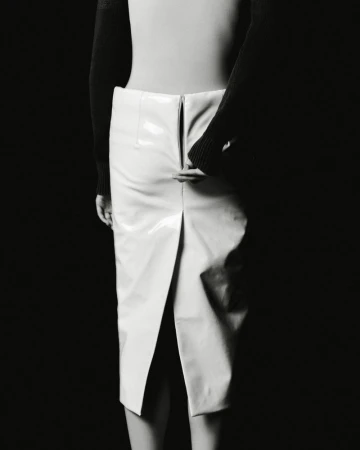 Model wearing a white glossy skirt and black top against a solid dark background for The Travel Almanac.