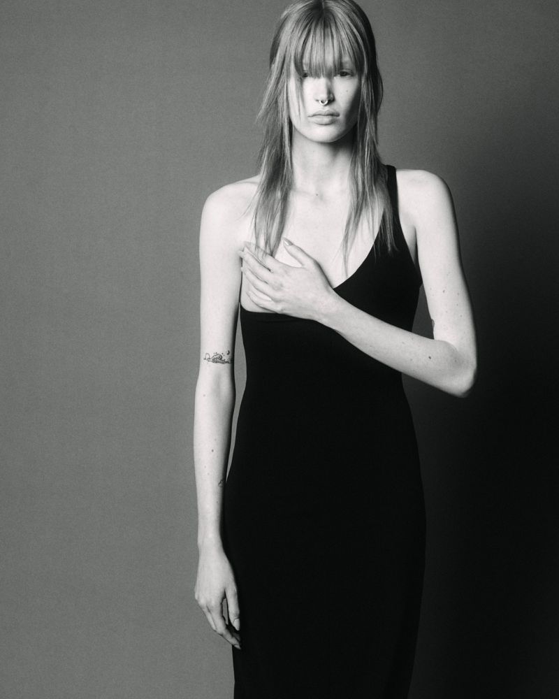 Monochrome portrait of a female model with bangs wearing a black dress against a plain grey studio backdrop.