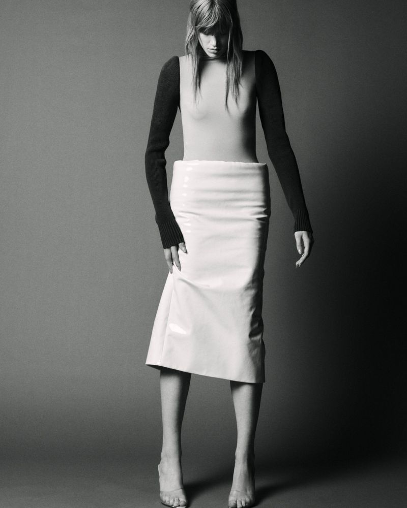 Model wearing a fitted top and white patent skirt against a grey studio backdrop