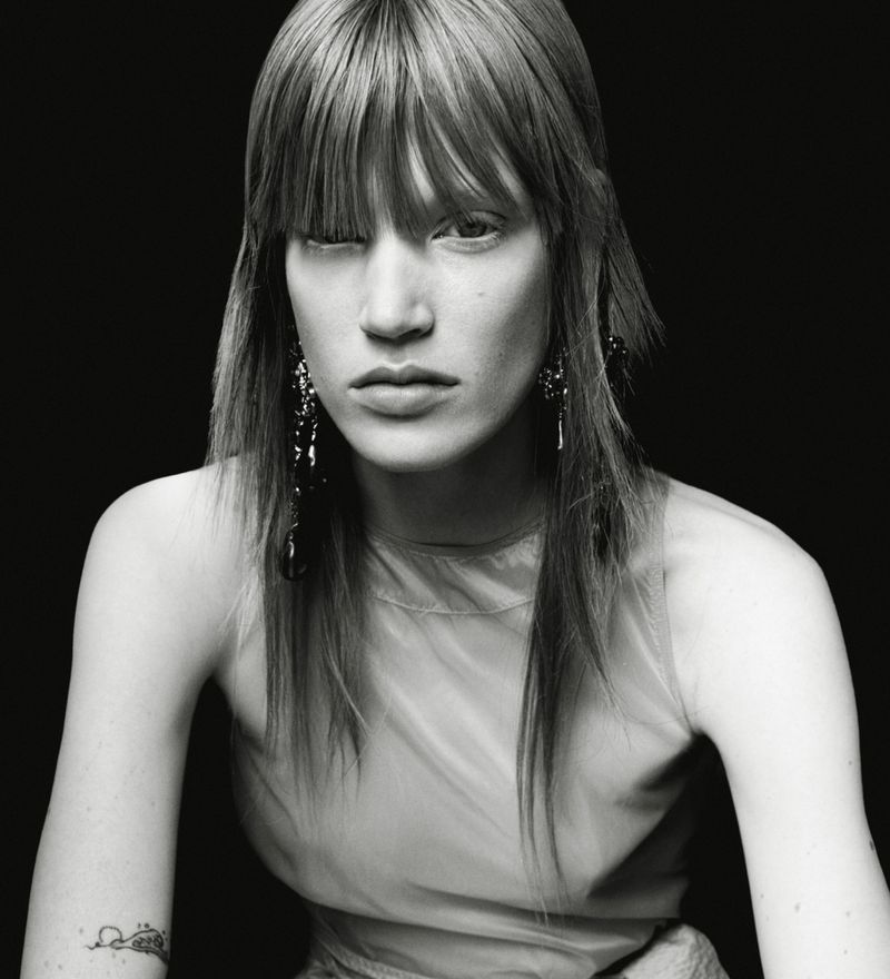 Monochrome portrait of a model with straight bangs and dark dangling earrings against a solid black background.