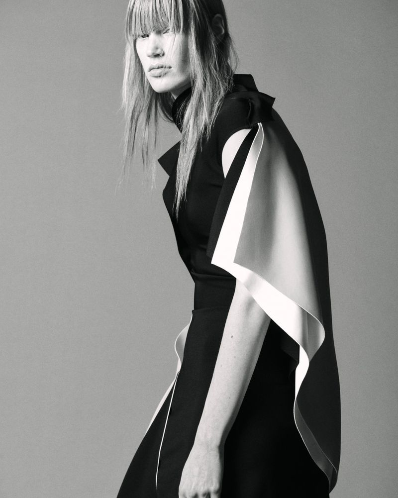 Model with fringe wearing a black dress with white trim against a plain grey studio backdrop