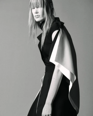 Model with fringe wearing a black dress with white trim against a plain grey studio backdrop