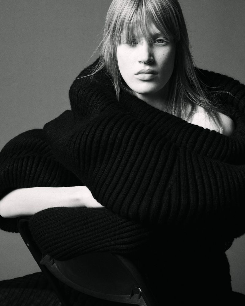 Model with long blonde hair and bangs wearing a bulky black knit sweater against a plain dark grey studio background.