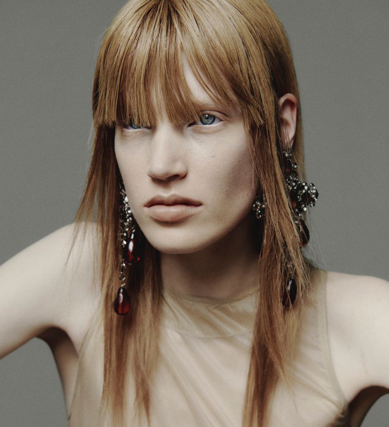 Close-up portrait of a model with blunt bangs wearing ornate drop earrings against a solid grey background.