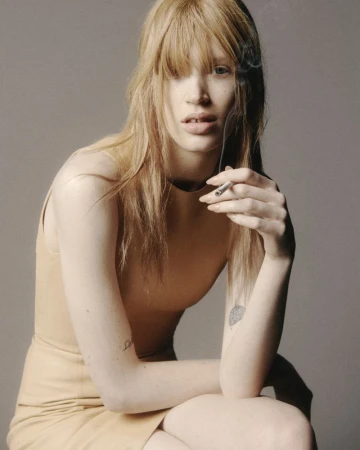 Close-up portrait of a blonde woman holding a cigarette against a plain grey studio backdrop