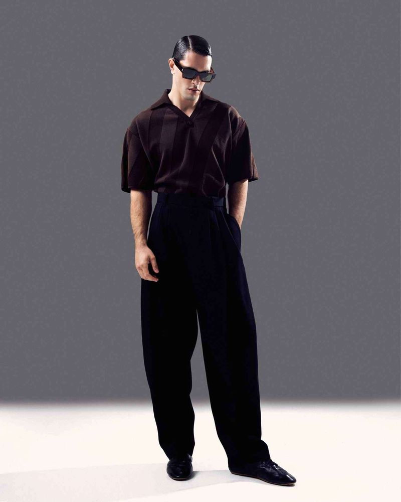 Full-length portrait of a man wearing dark sunglasses, brown polo shirt, and black trousers against a neutral grey studio backdrop.