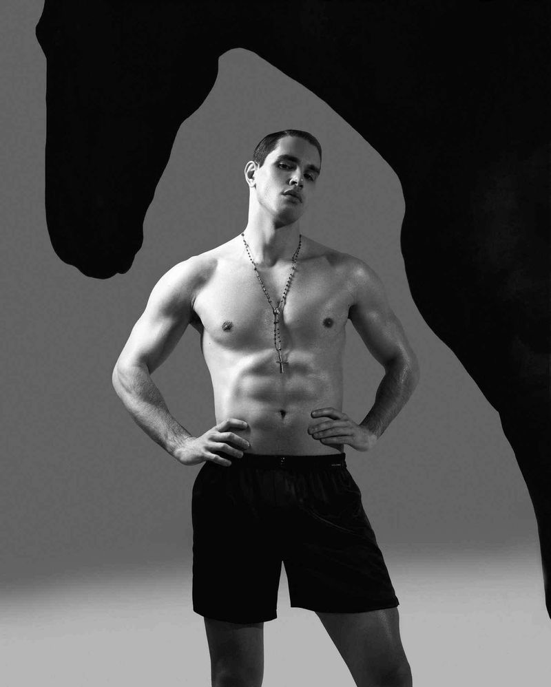 Shirtless male model wearing dark shorts and a necklace, posed against a grey background beneath a silhouette cutout of a horse head.