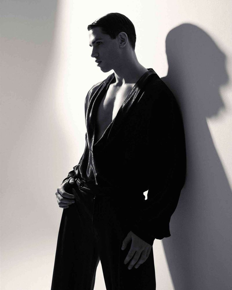 Portrait of a man in a black V-neck shirt leaning against a wall casting a shadow, for Glamour Man Bulgaria.