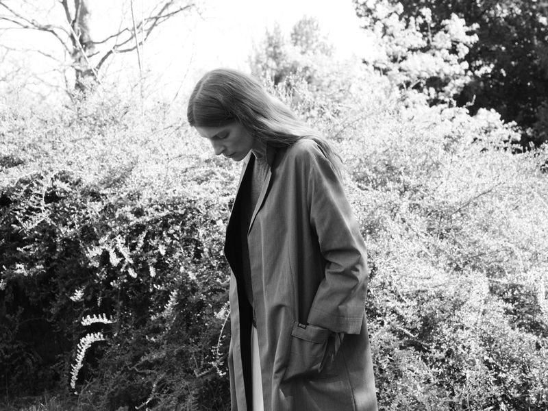Model with long hair wearing a belted coat standing outdoors surrounded by leafy bushes and dense trees.
