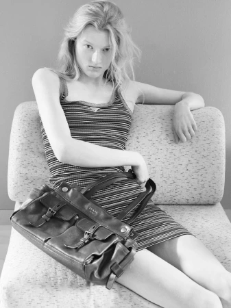 Model wearing a striped tank dress seated on a sofa while holding a dark leather handbag