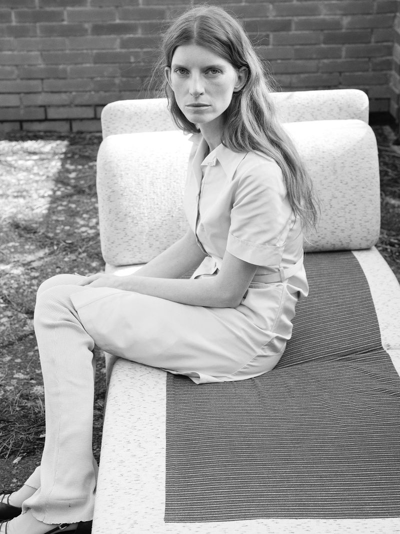 Monochrome shot of a woman with long hair, wearing a collared shirt and pants, sitting on an outdoor lounge chair in front of a brick wall.