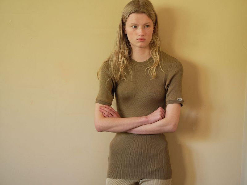 Model with long blonde hair wearing a brown ribbed t-shirt standing against a neutral yellow wall