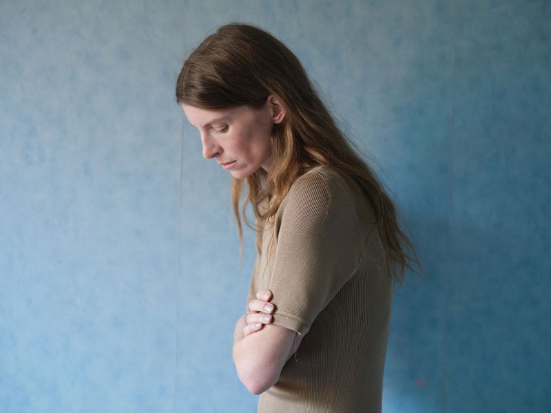 Profile portrait of a woman with long hair wearing a beige ribbed top standing against a textured blue wall.