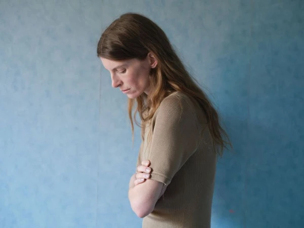 Profile portrait of a woman with long hair wearing a beige ribbed top standing against a textured blue wall.