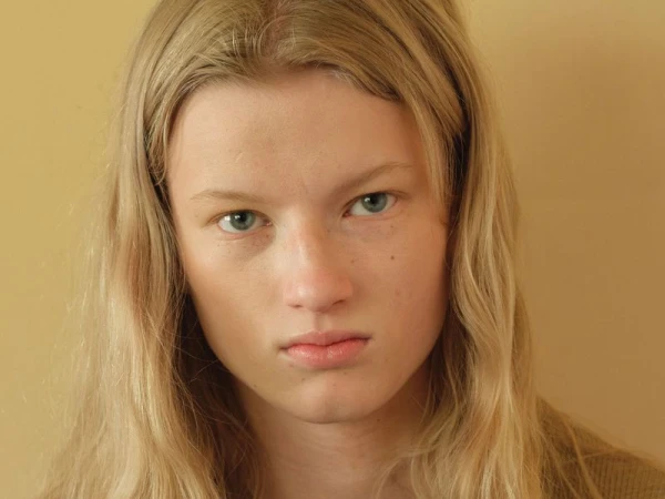 Close-up portrait of a young woman with long blonde hair and blue eyes against a neutral tan background.