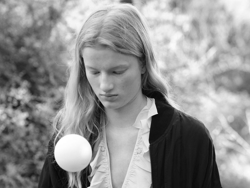 Monochrome portrait of a young woman with long blonde hair holding a white sphere in front of an outdoor background