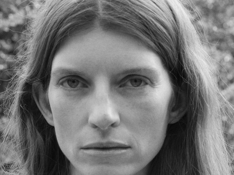 Close-up monochrome portrait of a woman with long hair looking directly into the camera lens against an out-of-focus outdoor backdrop.