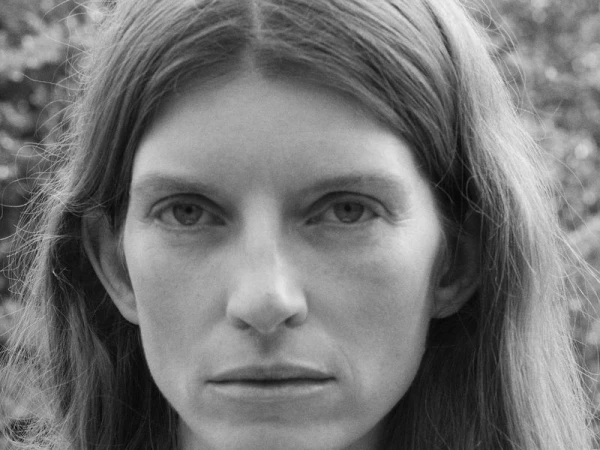 Close-up monochrome portrait of a woman with long hair looking directly into the camera lens against an out-of-focus outdoor backdrop.