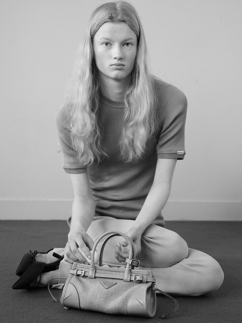 Model with long blonde hair seated on the floor wearing a fitted short-sleeve top and trousers, holding a handbag.