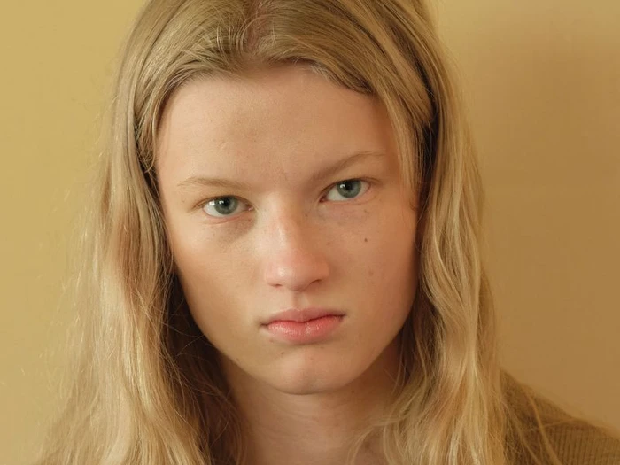 Close-up portrait of a young woman with long blonde hair and blue eyes against a neutral tan background.