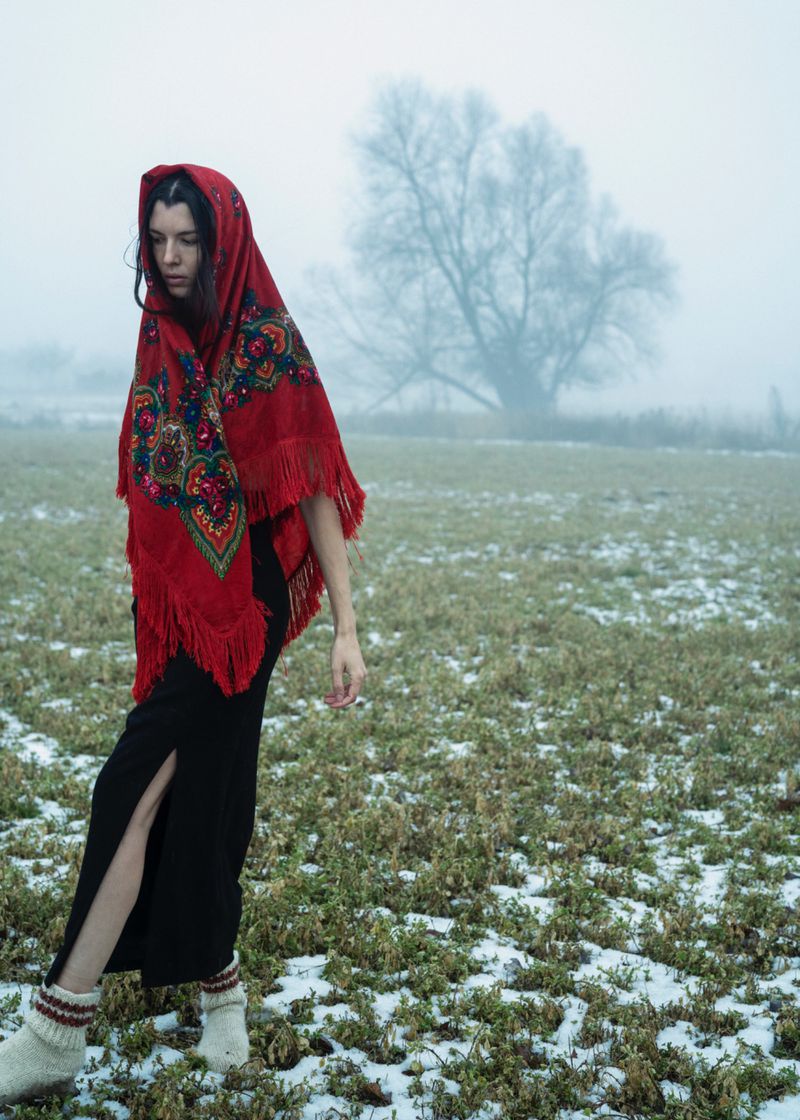 Woman wearing a bright red patterned headscarf and black skirt walking across a misty, snow-dusted field.