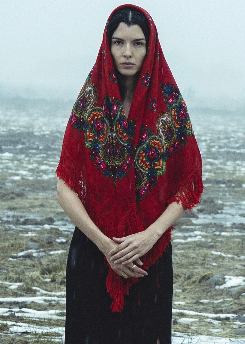 Woman wearing a bright red patterned headscarf and dark dress outdoors in a snowy landscape with mountains.