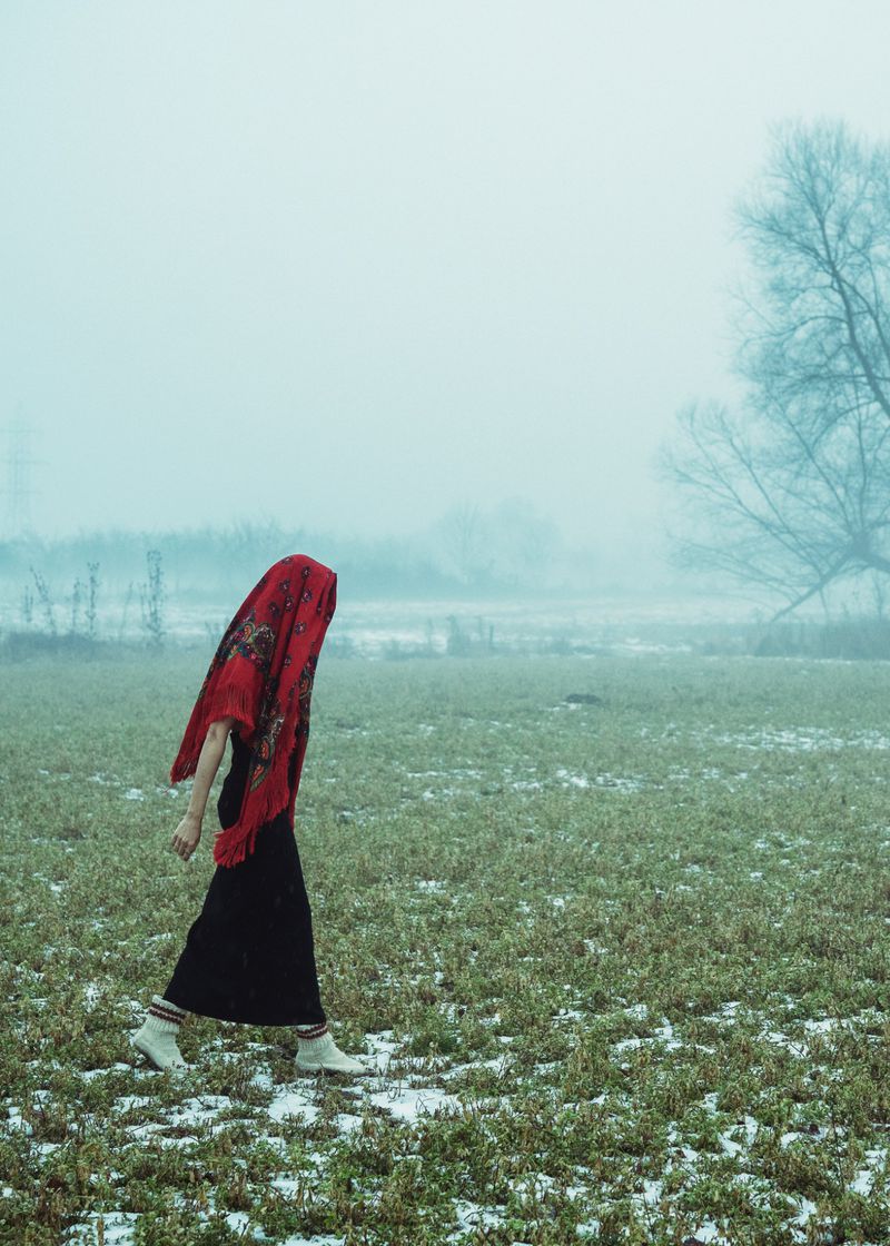 A person wearing a red patterned headscarf and dark skirt walks across a misty, sparse field under an overcast sky.