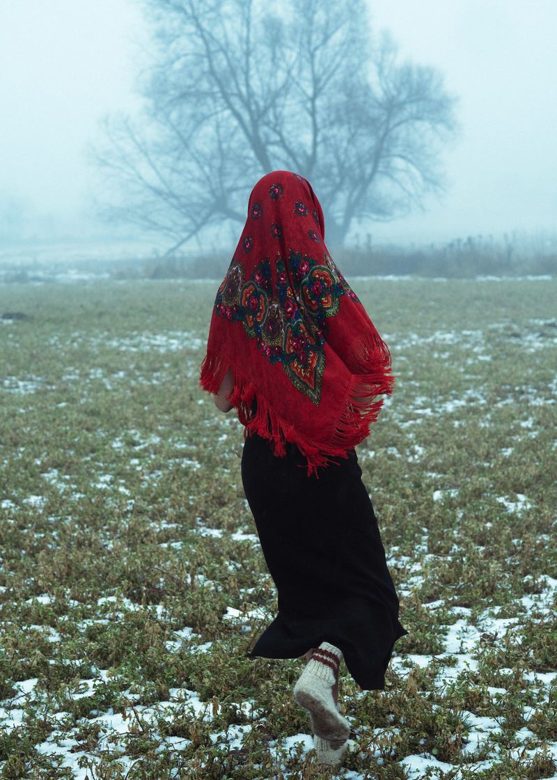 Figure in a red patterned headscarf and dark skirt running across a snowy field toward a bare tree in fog.