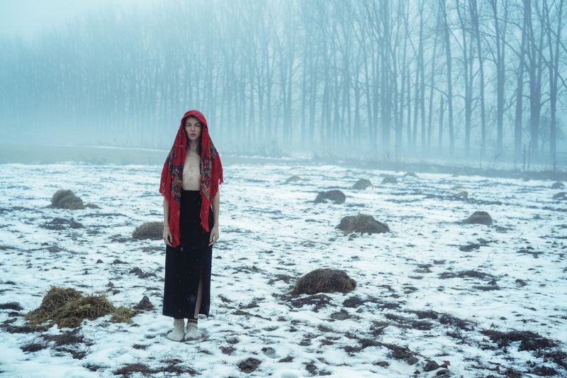 Woman wearing a red headscarf and dark skirt standing in a snowy field with bare trees in heavy fog