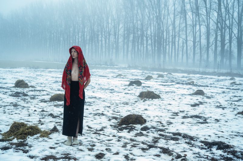 Model wearing a red headscarf and dark skirt standing in a snowy field with bare trees under a foggy sky.