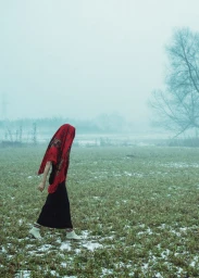 A person wearing a red patterned headscarf and dark skirt walks across a misty, sparse field under an overcast sky.